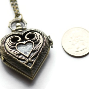 Puede incluir: Colgante con forma de coraz&oacute;n de color bronce antiguo con intrincados adornos y una peque&ntilde;a ventana transparente. El colgante est&aacute; en una cadena a juego. Una moneda est&aacute; al lado del colgante para comparar el tama&ntilde;o.