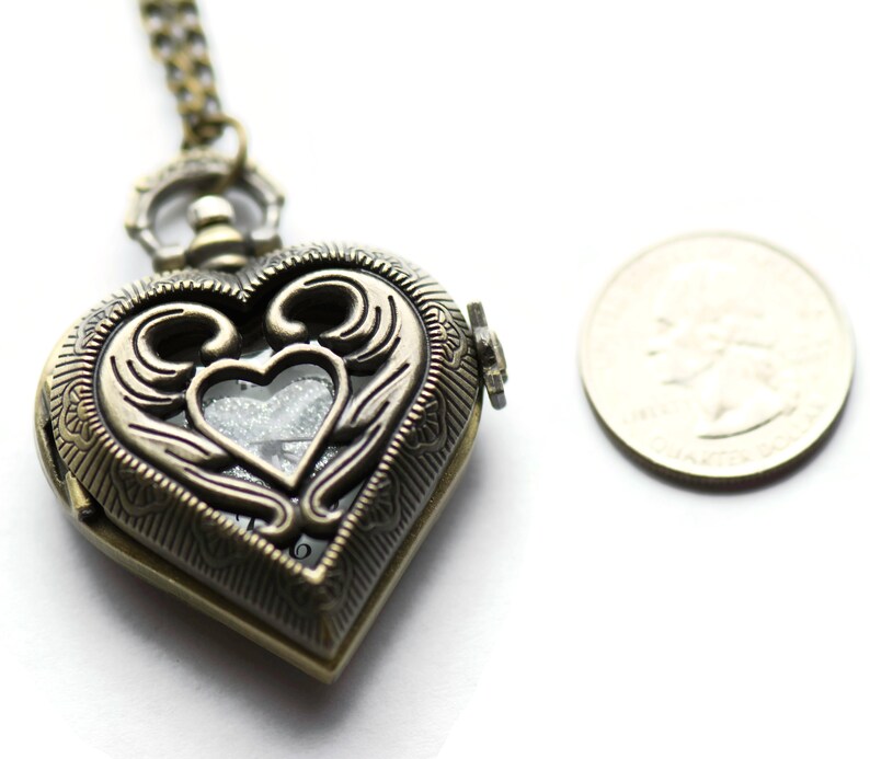 Puede incluir: Colgante con forma de coraz&oacute;n de color bronce antiguo con intrincados adornos y una peque&ntilde;a ventana transparente. El colgante est&aacute; en una cadena a juego. Una moneda est&aacute; al lado del colgante para comparar el tama&ntilde;o.