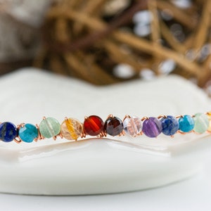 K&ouml;nnte beinhalten: Ein Regenbogen-Chakra-Armband aus Kupferdraht und verschiedenen Edelsteinen, darunter Amethyst, Lapislazuli, T&uuml;rkis, Aventurin, Citrin, Karneol, Granat, klarer Quarz und Amethyst.