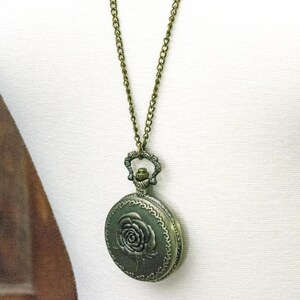 May include: Antique bronze-toned pendant necklace. The round pendant features a raised rose design on the front and hangs from a matching chain. The necklace is displayed on a white mannequin.