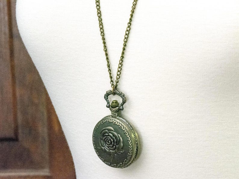 May include: Antique bronze-toned pendant necklace. The round pendant features a raised rose design on the front and hangs from a matching chain. The necklace is displayed on a white mannequin.