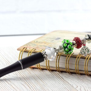 May include: A black pen with silver accents and a decorative charm. The charm includes a clear crystal, green polka dot bead, red bead, and a Christmas tree charm. The pen rests on a spiral-bound notebook.