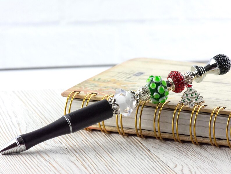 May include: A black pen with silver accents and a decorative charm. The charm includes a clear crystal, green polka dot bead, red bead, and a Christmas tree charm. The pen rests on a spiral-bound notebook.
