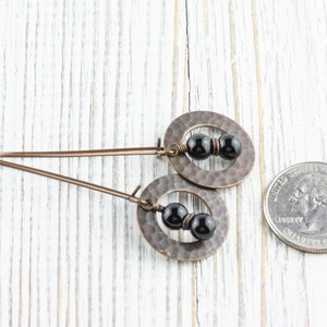 Peut inclure: Une paire de boucles d'oreilles cuivr&eacute;es avec une texture martel&eacute;e, chacune avec un design circulaire et trois perles noires. Les boucles d'oreilles sont pr&eacute;sent&eacute;es sur une surface en bois blanc &agrave; c&ocirc;t&eacute; d'une pi&egrave;ce de monnaie pour la r&eacute;f&eacute;rence de taille.