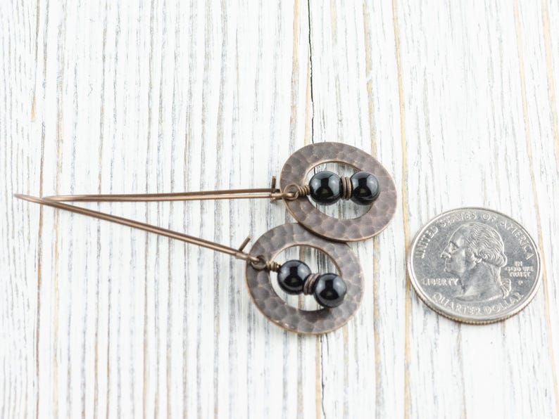 Peut inclure: Une paire de boucles d'oreilles cuivr&eacute;es avec une texture martel&eacute;e, chacune avec un design circulaire et trois perles noires. Les boucles d'oreilles sont pr&eacute;sent&eacute;es sur une surface en bois blanc &agrave; c&ocirc;t&eacute; d'une pi&egrave;ce de monnaie pour la r&eacute;f&eacute;rence de taille.