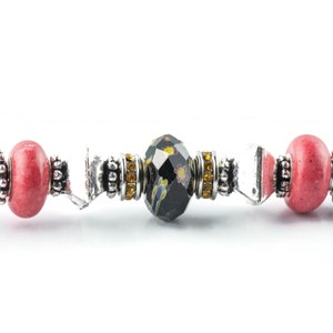 May include: Close-up of a decorative bottle stopper with a silver handle. The stopper features a central black faceted bead with a floral design, flanked by pink beads and silver accents. The design includes yellow and clear crystal accents.