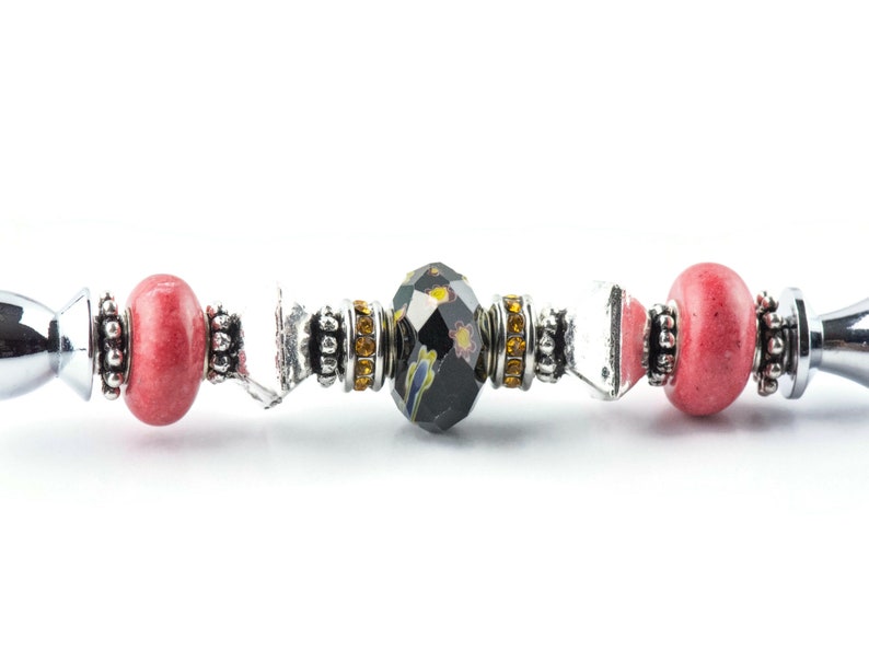 May include: Close-up of a decorative bottle stopper with a silver handle. The stopper features a central black faceted bead with a floral design, flanked by pink beads and silver accents. The design includes yellow and clear crystal accents.