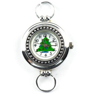 May include: A silver pocket watch with a white face and a green Christmas tree design. The watch has a silver chain and a small knob on the side.