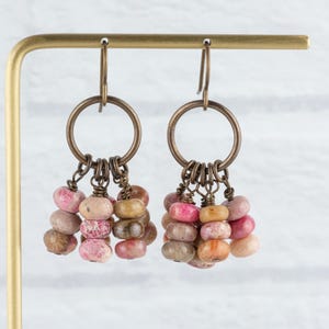 Op de afbeelding: Een paar oorhangers met bronzen ringen en haken. Elke oorbel heeft een cluster van kleine, kleurrijke, afgeronde kralen in roze, bruin en beige, die aan de ring hangen. De oorbellen worden getoond op een goudkleurige standaard.