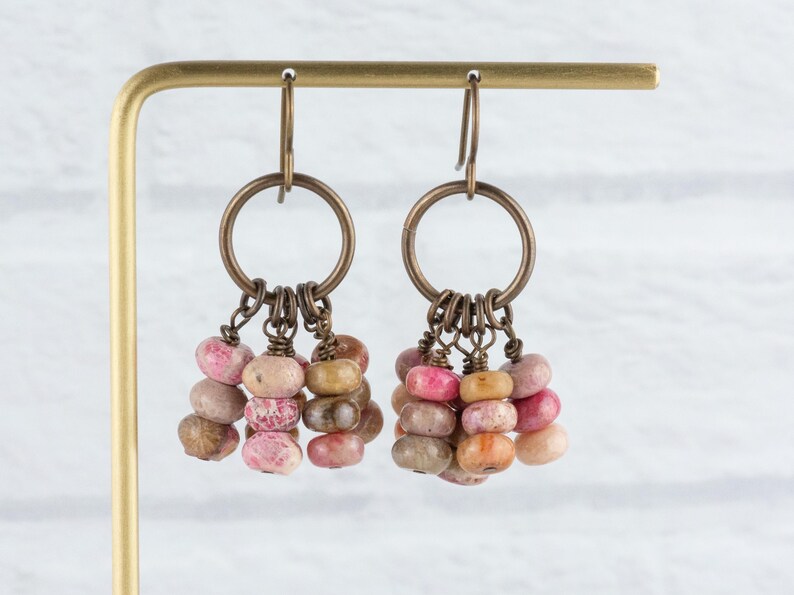 Op de afbeelding: Een paar oorhangers met bronzen ringen en haken. Elke oorbel heeft een cluster van kleine, kleurrijke, afgeronde kralen in roze, bruin en beige, die aan de ring hangen. De oorbellen worden getoond op een goudkleurige standaard.
