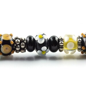May include: A close-up of a bracelet with black, white, and yellow beads. The beads are arranged in a pattern and have a variety of shapes and sizes. The bracelet is made of glass and metal.