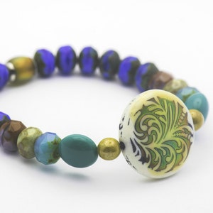 Puede incluir: Un brazalete de cuentas azules, marrones, verdes y doradas con un dije de cer&aacute;mica blanca con un dise&ntilde;o floral verde.