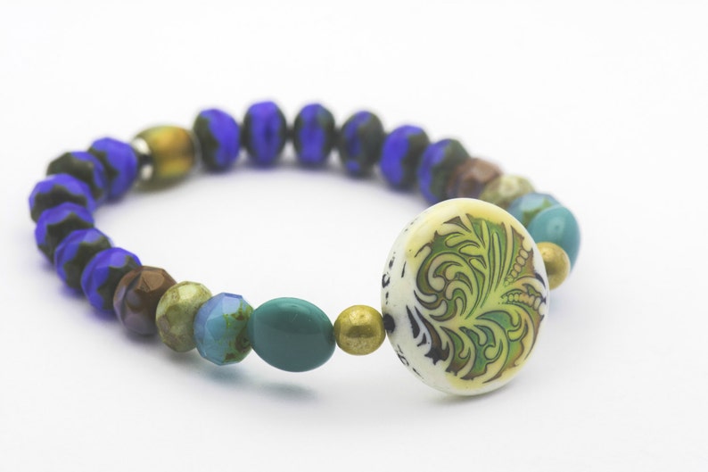 Puede incluir: Un brazalete de cuentas azules, marrones, verdes y doradas con un dije de cer&aacute;mica blanca con un dise&ntilde;o floral verde.