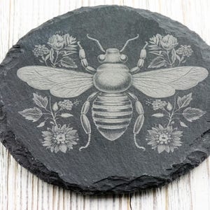 Op de afbeelding: Een zwarte leisteen onderzetter met een wit gegraveerd beeld van een bij omringd door bloemen.