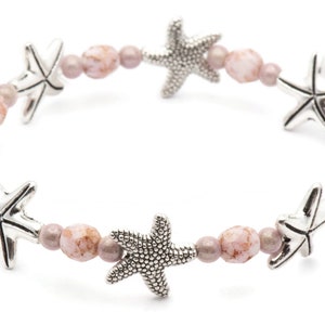K&ouml;nnte beinhalten: Ein Stretch-Armband mit silbernen Seestern-Anh&auml;ngern und rosa- und malvenfarbenen Perlen. Das Armband hat ein Strandthema und ist f&uuml;r den t&auml;glichen Gebrauch geeignet. Ein sch&ouml;nes Accessoire.