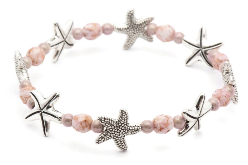 K&ouml;nnte beinhalten: Ein Stretch-Armband mit silbernen Seestern-Anh&auml;ngern und rosa- und malvenfarbenen Perlen. Das Armband hat ein Strandthema und ist f&uuml;r den t&auml;glichen Gebrauch geeignet. Ein sch&ouml;nes Accessoire.
