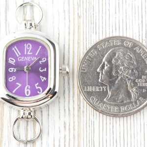 May include: A silver-toned Geneva watch with a purple face and white numerals. The watch is rectangular with rounded corners and has a small crown. A U.S. quarter is next to the watch for size comparison.