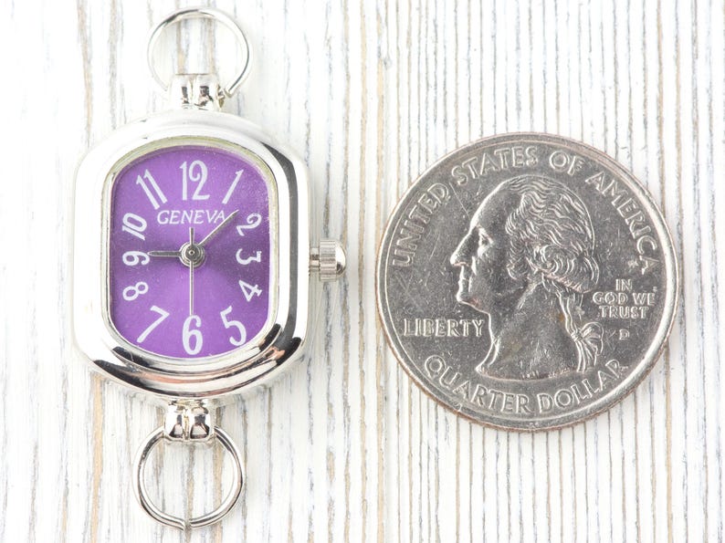May include: A silver-toned Geneva watch with a purple face and white numerals. The watch is rectangular with rounded corners and has a small crown. A U.S. quarter is next to the watch for size comparison.