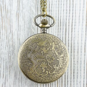 K&ouml;nnte beinhalten: Antike Taschenuhr aus Messing mit einem verzierten floralen Design auf dem Deckel. Die Uhr h&auml;ngt an einer passenden Kette. Der Hintergrund ist eine wei&szlig; lackierte Holzoberfl&auml;che.
