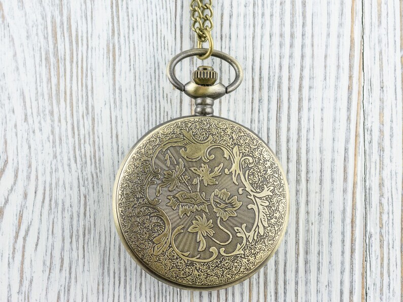 K&ouml;nnte beinhalten: Antike Taschenuhr aus Messing mit einem verzierten floralen Design auf dem Deckel. Die Uhr h&auml;ngt an einer passenden Kette. Der Hintergrund ist eine wei&szlig; lackierte Holzoberfl&auml;che.