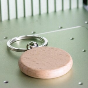 Op de afbeelding: Een houten sleutelhanger met een ronde, lichtbruine schijf bevestigd aan een zilveren sleutelring.