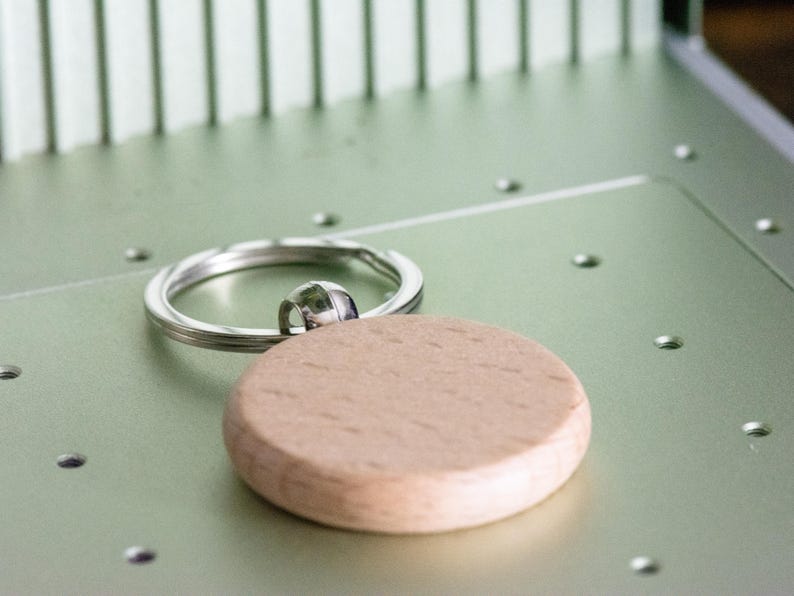 Op de afbeelding: Een houten sleutelhanger met een ronde, lichtbruine schijf bevestigd aan een zilveren sleutelring.
