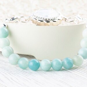 May include: A light blue gemstone bracelet with a silver clasp and a silver charm. The charm has a white stone in the center and a decorative silver border.