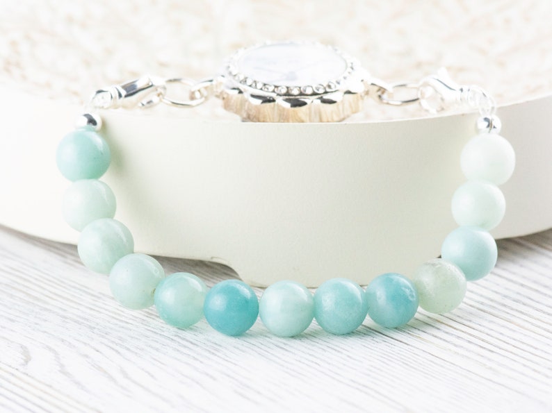 May include: A light blue gemstone bracelet with a silver clasp and a silver charm. The charm has a white stone in the center and a decorative silver border.