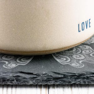 May include: A beige ceramic mug with the word "LOVE" printed in blue on the side. The mug is sitting on a black slate coaster with a decorative design.