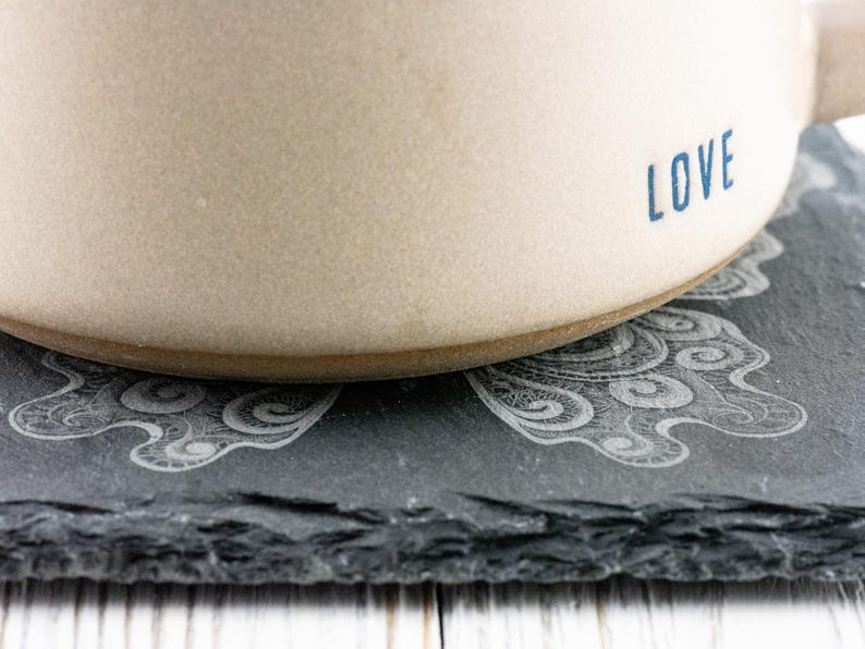 May include: A beige ceramic mug with the word "LOVE" printed in blue on the side. The mug is sitting on a black slate coaster with a decorative design.