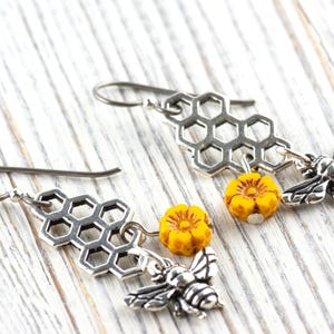 May include: Silver-tone earrings with a honeycomb design, yellow flower beads, and bee charms. The earrings are displayed on a white wooden surface, highlighting the intricate details of the jewellery.