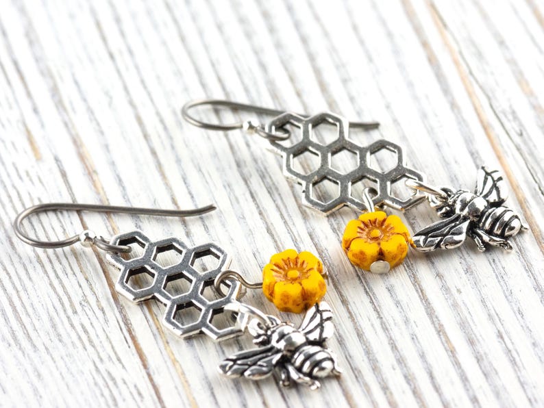 May include: Silver-tone earrings with a honeycomb design, yellow flower beads, and bee charms. The earrings are displayed on a white wooden surface, highlighting the intricate details of the jewellery.