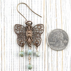 May include: A bronze-colored butterfly earring with intricate filigree wings and three dangling, pale green beads. The earring is displayed on a white wooden surface next to a quarter for size comparison.