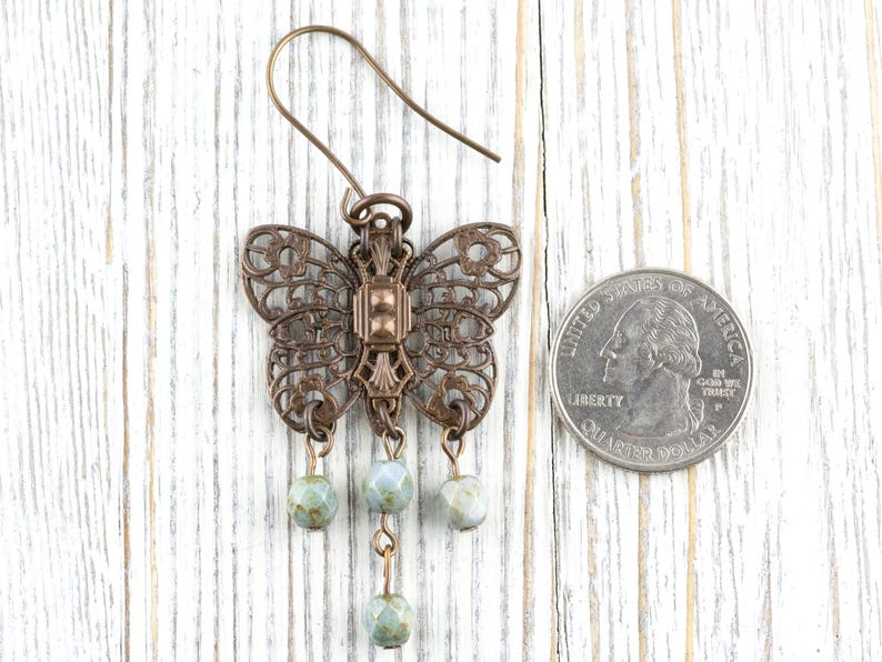 May include: A bronze-colored butterfly earring with intricate filigree wings and three dangling, pale green beads. The earring is displayed on a white wooden surface next to a quarter for size comparison.
