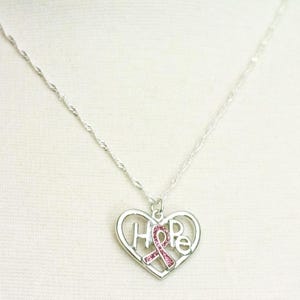 Pode incluir: Um colar prateado com um pingente em forma de cora&ccedil;&atilde;o. O pingente apresenta a palavra "HOPE" e uma fita rosa, simbolizando a conscientiza&ccedil;&atilde;o sobre o c&acirc;ncer de mama. O colar tem uma corrente delicada.