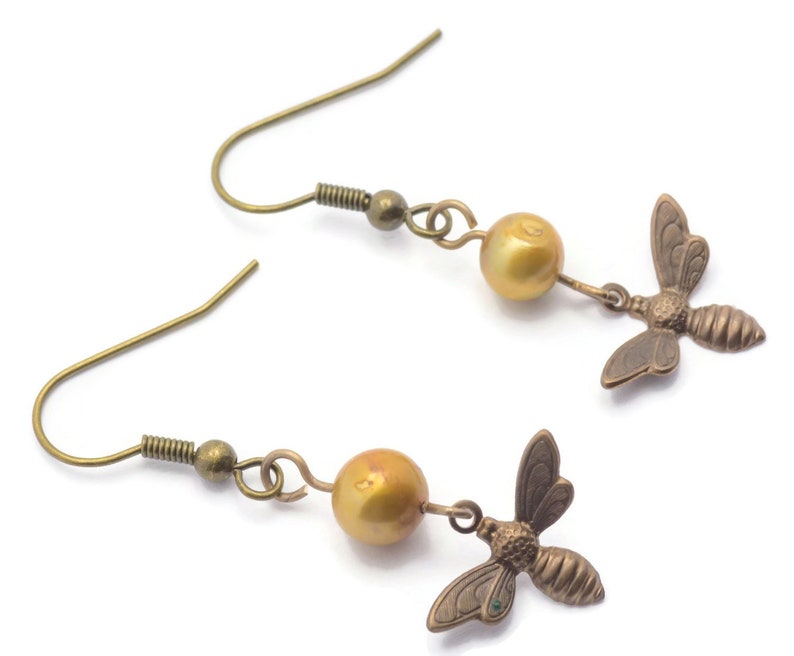 May include: A pair of gold-toned earrings featuring a small bee charm and a golden pearl bead on each earring.