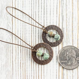 Puede incluir: Un par de pendientes de color cobre con textura martillada, cada uno con un dise&ntilde;o circular y dos cuentas verdes y marrones. Los pendientes se muestran junto a una moneda de veinticinco centavos estadounidense.