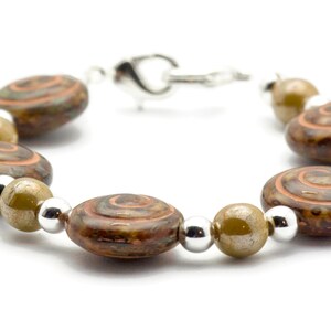 May include: A bracelet with brown, oval beads with a spiral pattern in brown and orange. The bracelet also has silver and olive-green beads, connected by a silver clasp. Jewellery.