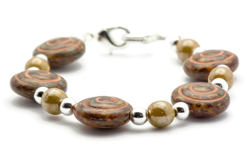 May include: A bracelet with brown, oval beads with a spiral pattern in brown and orange. The bracelet also has silver and olive-green beads, connected by a silver clasp. Jewellery.