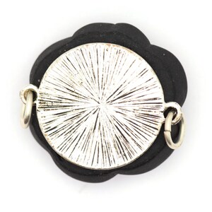 May include: A silver-toned pendant with a circular design and a black flower-shaped backing. The pendant has two silver jump rings for attaching to a chain.