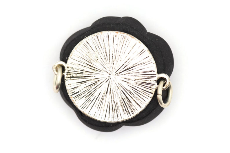 May include: A silver-toned pendant with a circular design and a black flower-shaped backing. The pendant has two silver jump rings for attaching to a chain.