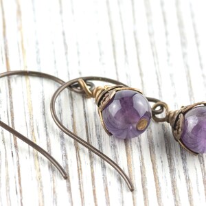 K&ouml;nnte beinhalten: Ein Paar handgefertigte Ohrringe mit polierten, runden, lila Amethystperlen. Die Perlen sind in aufwendigen Metallarbeiten im Antik-Stil mit Bronze-Finish eingefasst. Die Ohrringe haben einfache, gebogene Ohrhaken.