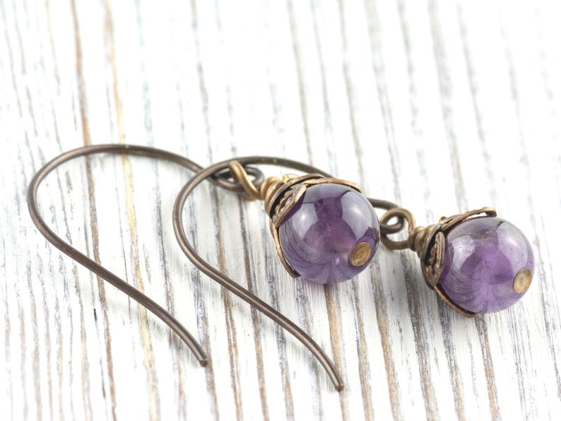 K&ouml;nnte beinhalten: Ein Paar handgefertigte Ohrringe mit polierten, runden, lila Amethystperlen. Die Perlen sind in aufwendigen Metallarbeiten im Antik-Stil mit Bronze-Finish eingefasst. Die Ohrringe haben einfache, gebogene Ohrhaken.