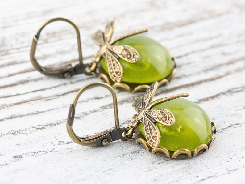 May include: A pair of green glass earrings with dragonfly accents. The earrings have a bronze finish and are designed with a hook closure.