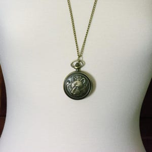 May include: A gold chain necklace with a round, antique-style pocket watch pendant featuring an embossed wolf head design.