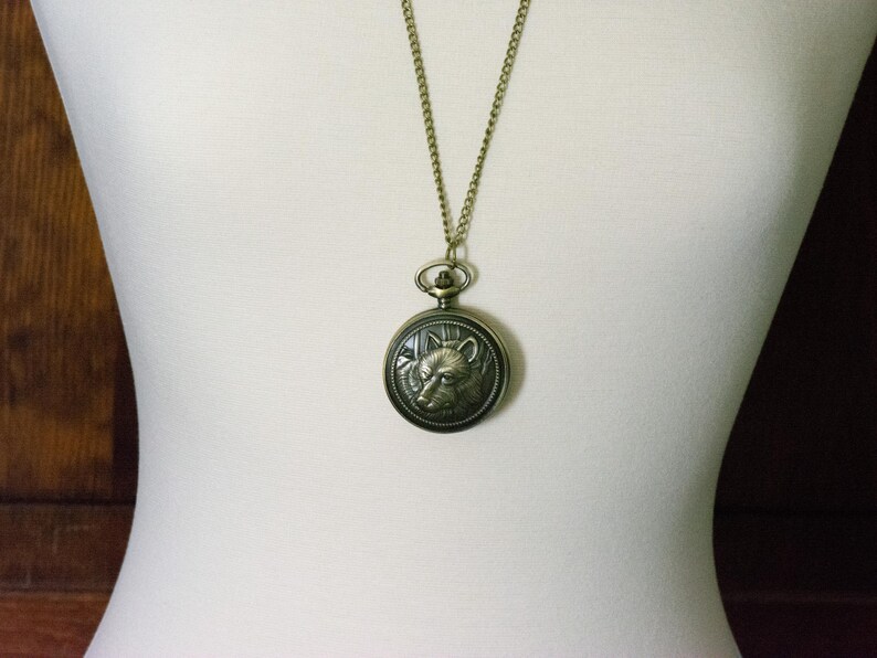 May include: A gold chain necklace with a round, antique-style pocket watch pendant featuring an embossed wolf head design.