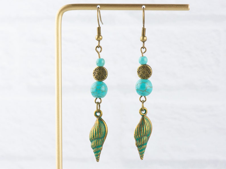 K&ouml;nnte beinhalten: Ein Paar t&uuml;rkisfarbene und bronzene Ohrringe mit einem Muschel-Anh&auml;nger. Die Ohrringe bestehen aus einer t&uuml;rkisfarbenen Perle, einer bronzenen Scheibe mit einem komplizierten Design und einer t&uuml;rkisfarbenen Perle. Der Muschel-Anh&auml;nger ist bronzefarben und hat ein detailliertes Design.