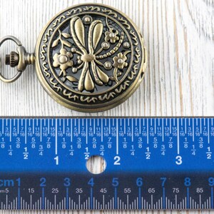 May include: Antique bronze pocket watch with a floral and dragonfly design. The watch is approximately 2 inches in diameter. A blue ruler with centimeter and millimeter markings is placed next to the watch.