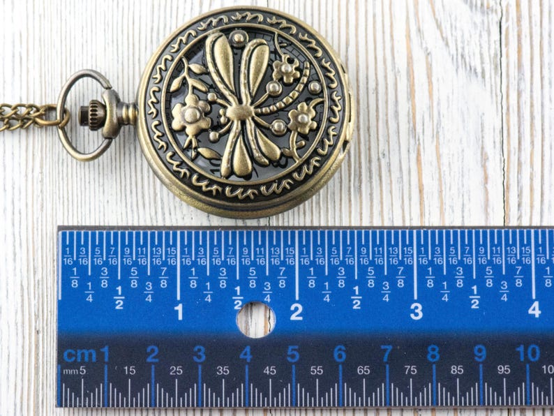 May include: Antique bronze pocket watch with a floral and dragonfly design. The watch is approximately 2 inches in diameter. A blue ruler with centimeter and millimeter markings is placed next to the watch.