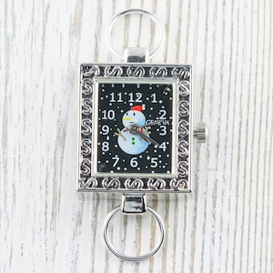 K&ouml;nnte beinhalten: Eine silberne Taschenuhr mit schwarzem Zifferblatt und einem Schneemann-Motiv. Die Uhr hat einen Schl&uuml;sselring.